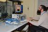 Man with beard and glasses in light grey sweater sitting on the right looking at computer screen on the left, with mass of blue wires in the foreground.