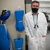 A man in a white lab coat and black face mask (Jeffrey Herrera) stands near a wall of protective equipment like long gloves.
