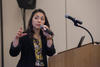 A woman (Danielle Santos) stands at a podium holding a microphone and gesturing with her other hand. 