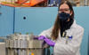 masked woman with glasses, purple gloves, and a labcoat stands next to a large aluminum sample chamber