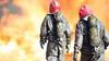 Fire scene with two individuals appearing to be firefighters facing the fire. Individuals are wearing red helmets, camo gear, gloves, and air tanks.