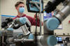 A man wearing a face mask touches a computer screen in a lab with robotic arms in the foreground.