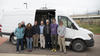 Eight adults pose for a photo standing by a white van with the side door open, showing equipment inside. 