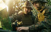 Camouflaged military officer using a radio and laptop giving orders to other soldiers in a tent in the forest.