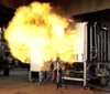 A fireball rushes out of the opening of a small metal compartment in a concrete room. 