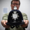 A blurry figure in the background holds out a black police helmet with a silver seal that reads "Norfolk Constabulary."