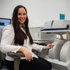Ileana Pazos sits and smiles for a photo next to scientific equipment. She's holding tweezers next to the machine.