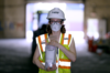 A woman in a NIST hard hat and other safety gear stands inside a warehouse holding a plastic bag labeled "Evidence."