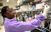 Alshae' Logan-Jackson wears a purple coverall and safety glasses in the lab as she uses tweezers to hold a small white disk. 