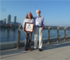Ursula Kattner and guest pose for a photo with her Gibbs Triangle Award in front of the water.