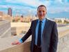 Robert DeJaco poses outdoors with a city skyline and blue sky behind him. 