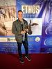 Man in black jeans, black shoes, plaid shirt and khaki jacket stands in front of a banner that says Ethos Film Awards holding an award