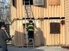 A firefighter climbs a ladder into a shipping container, while a person to their left tracks their location.