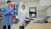 Ryan Falkenstein-Smith and Amy Mensch, both wearing lab coats and gloves, stand in the lab next to a black camera-like device on a table. 