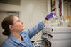 Alix Rodowa wears safety glasses and gloves in the lab as she reaches for a container of clear liquid on top of a piece of scientific equipment. 