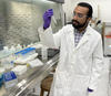 George Caceres wears safety glasses and a lab coat as he looks at a plastic test tube he is holding.