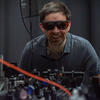 Krister Shalm, wearing safety glasses, smiles over a lab table covered with electronics equipment.