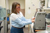 A researcher wearing safety glasses stands in the lab, reaching into a metal device on the lab table. 