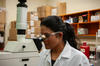 A woman wearing a lab coat and safety glasses stares into a large microscope.
