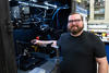 A bearded man poses smiling next to an open cabinet of equipment in a lab. 