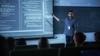 A young male teacher stands at the front of a darkened classroom, pointing at computer code on a large screen. 