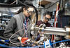 Two researchers wearing safety glasses are seen standing in the lab surrounded by wiring and electronics.