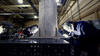 Two people wearing welding masks and other safety equipment bend over a table to work on a large metal cylinder.
