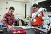 Two young men stand a lab table fitted with a white articulated robot arm.