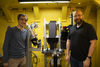 Cyrus Daugherty and Jake LaManna, wearing safety glasses, pose on either side of an imaging device holding a replica scroll.