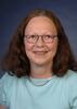 Ursula Kattner, a woman with long brown hair and wearing glasses, smiling for a professional headshot in front of a studio backdrop.