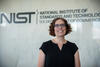 A woman with short curly brown hair and glasses smiles at the camera. NIST logo is in the background. 