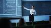 A female instructor wearing glasses presents code on a large screen to a classroom.